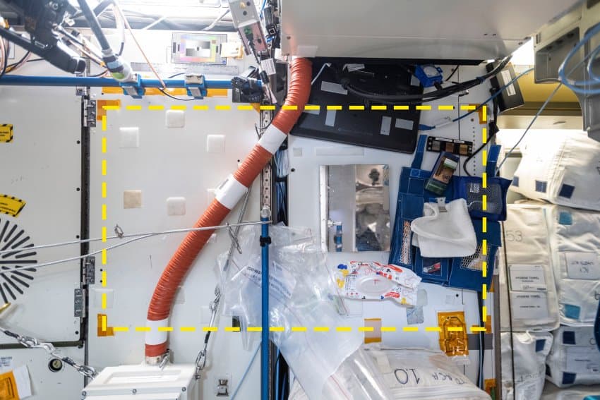 A photograph of a section of wall on the iss, it shows various ducting and plastic bags and an open toiletries bag with mirror, towel and assorted other personal hygiene items