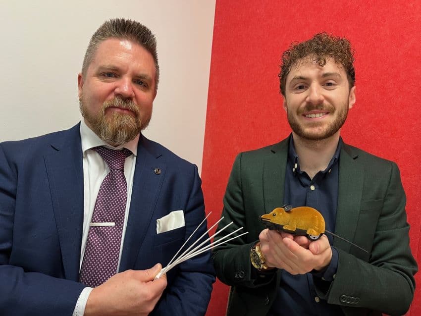 Two people smiling at camera, one holding long white whiskers and one holding toy rat robot