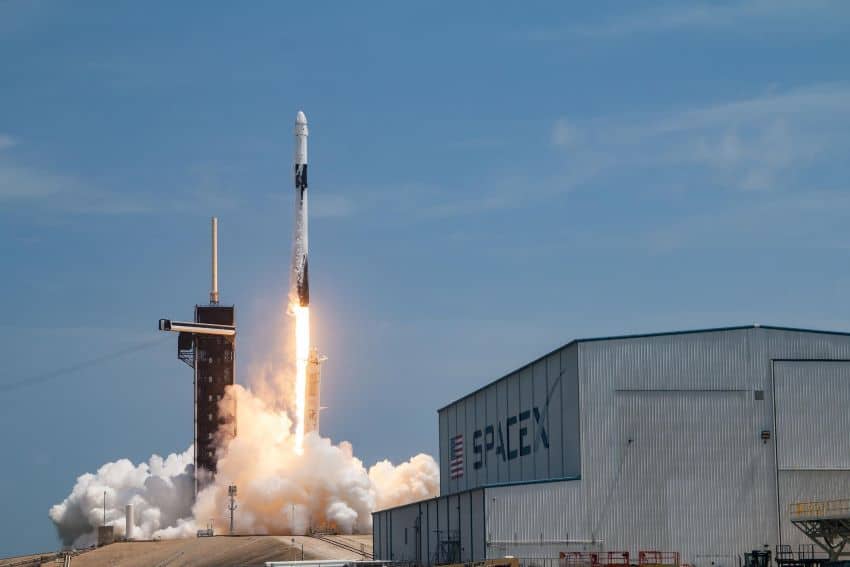 Spacex rocket launches off pad with spacex warehouse in foreground