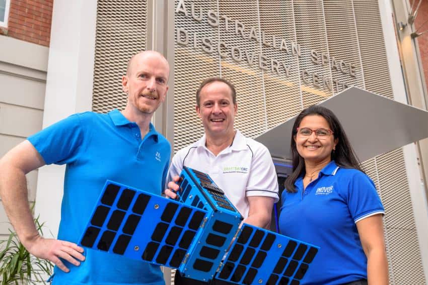 A photograph of three people, two men and a woman, holding a life-sized blue model of satellite kanyini.
