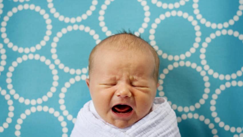 A newborn baby crying. It is swaddled in a white blanket and placed on a blue and white blanket