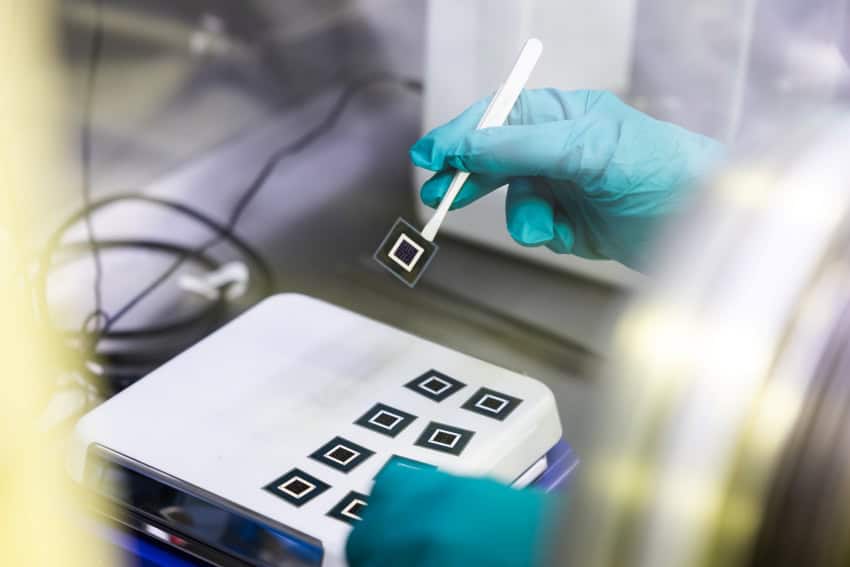 A gloved hand holds a small square solar cell with a set of tweazers