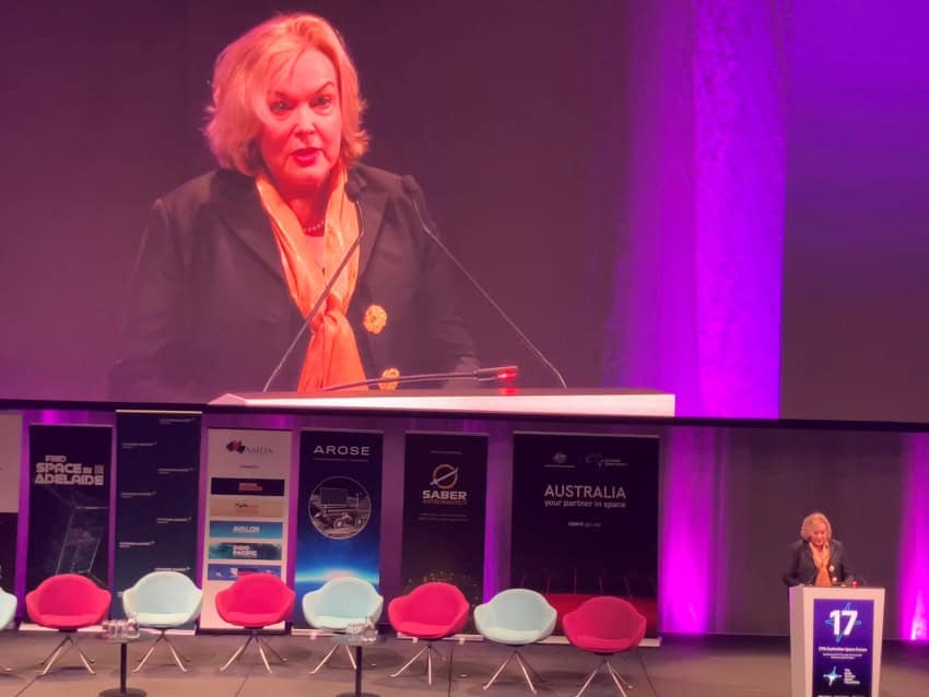 A photograph of a woman with short blonde hair standing at a podium during a speech.