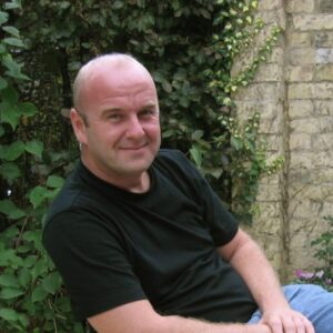 A photograph of a man wearing a black tshirt and blue jeans. He is sitting outside in front of some greenery