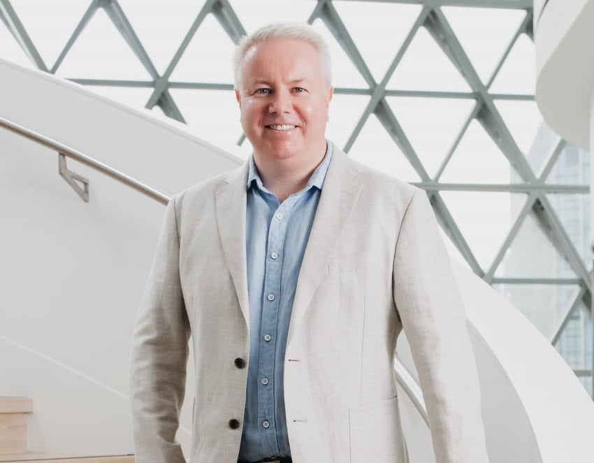 A man with short white hair, wearing a pale blue shirt and off-white linen blazer, smiles at the camera, he is standing in front of a white staircase and wall of triangular windows.