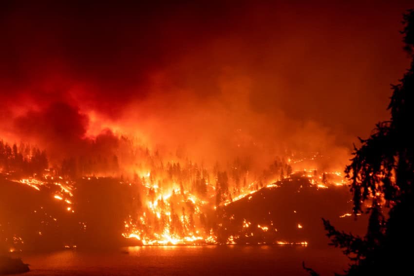 Photograph of a fores-covered hillside at night. The hill is glowing orange with the light and smoke of wildfires.