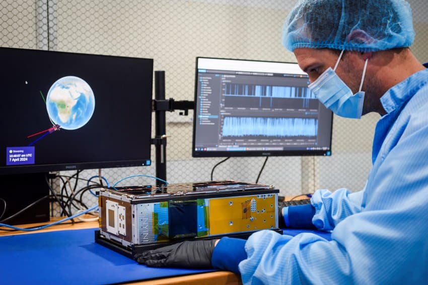 A photograph of a person wearing ppe, including lab coat, gloves and mask handling a small cereal box-sized satellite.