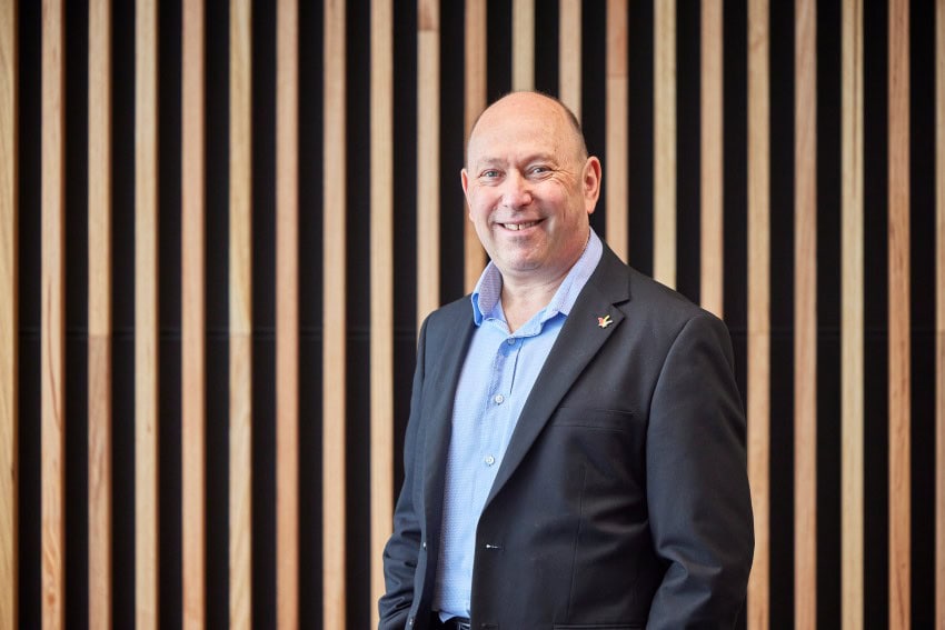 Photograph of a man wearing a charcoal grey suit and blue shirt. He is standing in front of a wooden panelled background