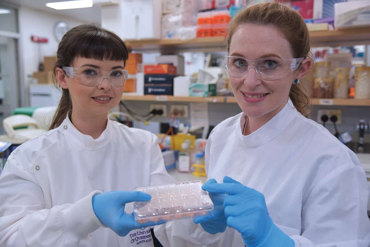 Two women in white lab coats wearing blue gloves, holding an plastic dish with several compartments.