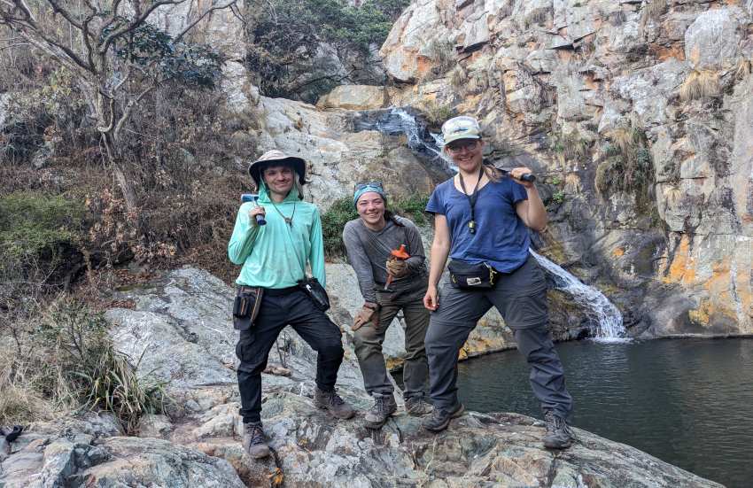 Three scientists stand in the field, near a rocky outcrop and a body of water.