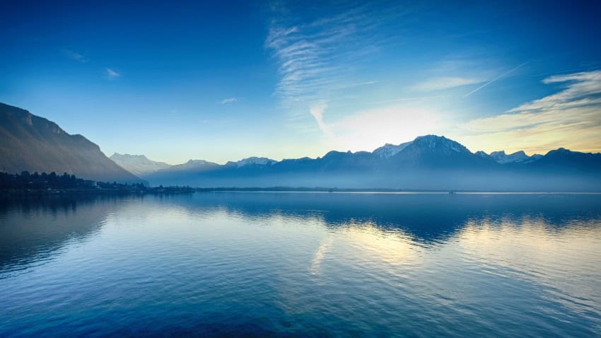 A photograph of a still blue lake with the swiss alphs in the background.