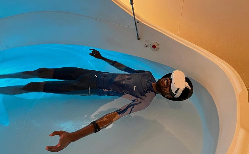Photograph of a man wearing swimwear floating in a flotation tank lit by blue light while wearing vr goggles
