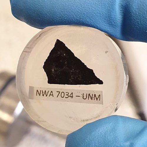 Blue glove fingers holding black sample of rock in dish