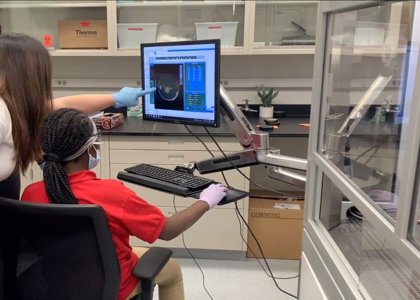 A photograph of a young child wearing a red polo shirt sitting at a computer in a laboratory with an adult pointing at a screen