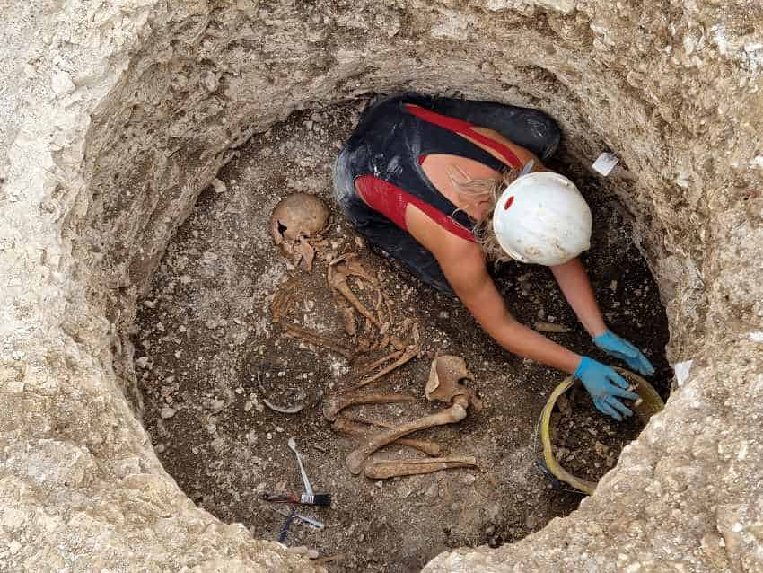 Photograph of a woman removing rock and soil from around a skeleton at the bottom of a circular hole.