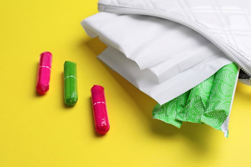 Assorted period products, including pads and tampons, spilling out of a small white bag, against a yellow background