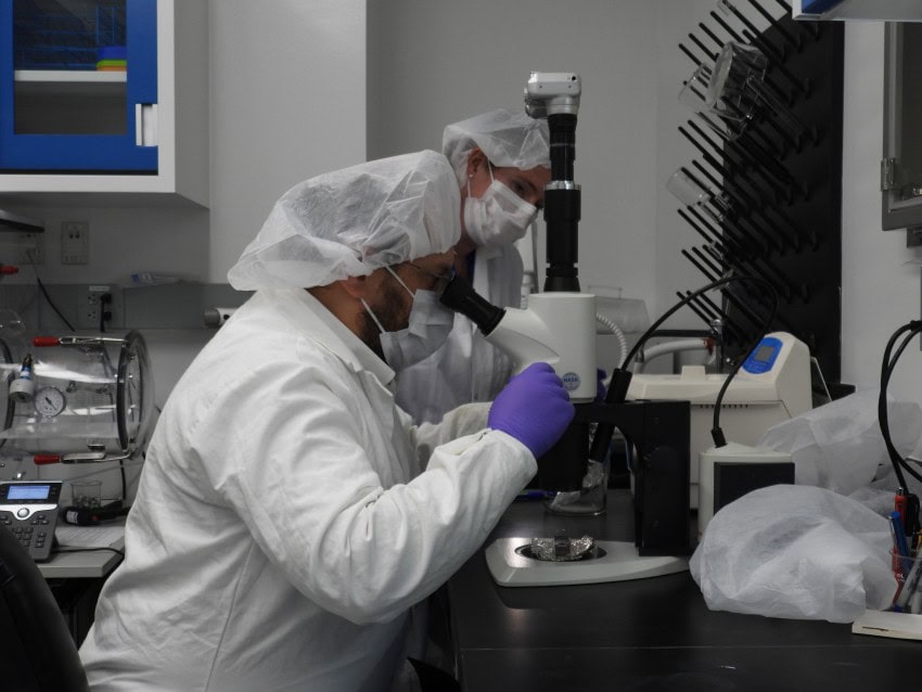 A man in a white lab coat peers into a microscope that is looking at samples from bennu.