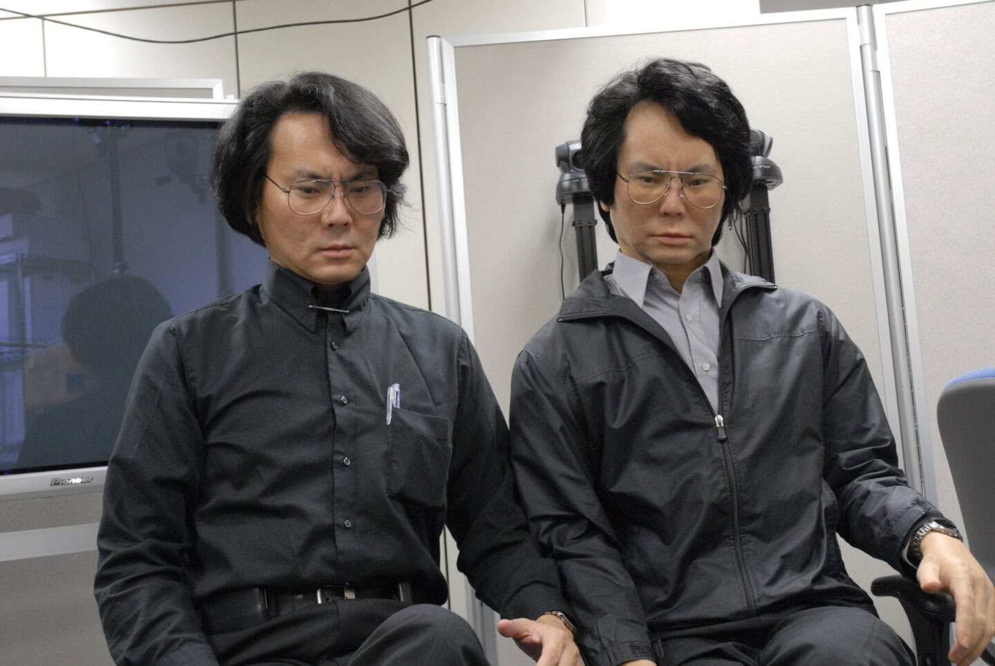 Japanese man wearing black clothes and glasses next to a robot made in his image.