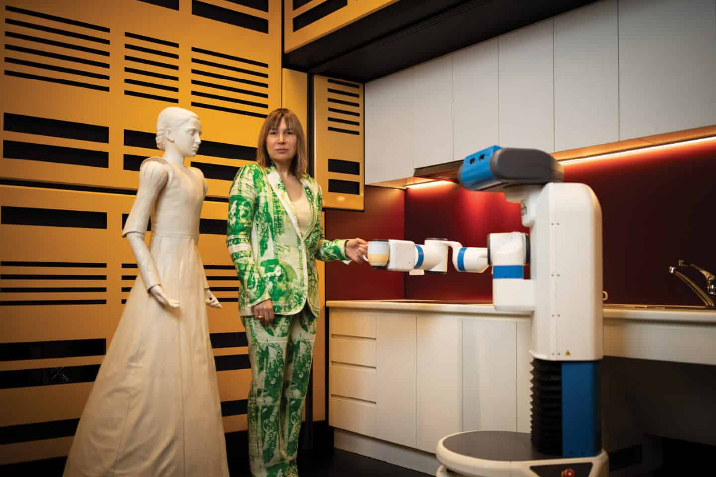 Woman in green and white print suit holding a mug held by a robot, next to a white robot that looks like a woman.