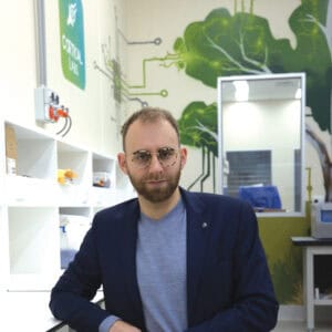 Man in t-shirt and suit jacket with glasses sitting in a lab.