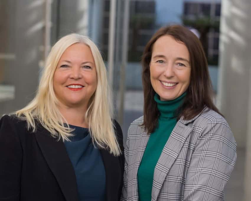 2 women smile at the camera. The one on the length had mid-length straight blond hair and the one on the right has straight brown hair. Both are wearing blazers.