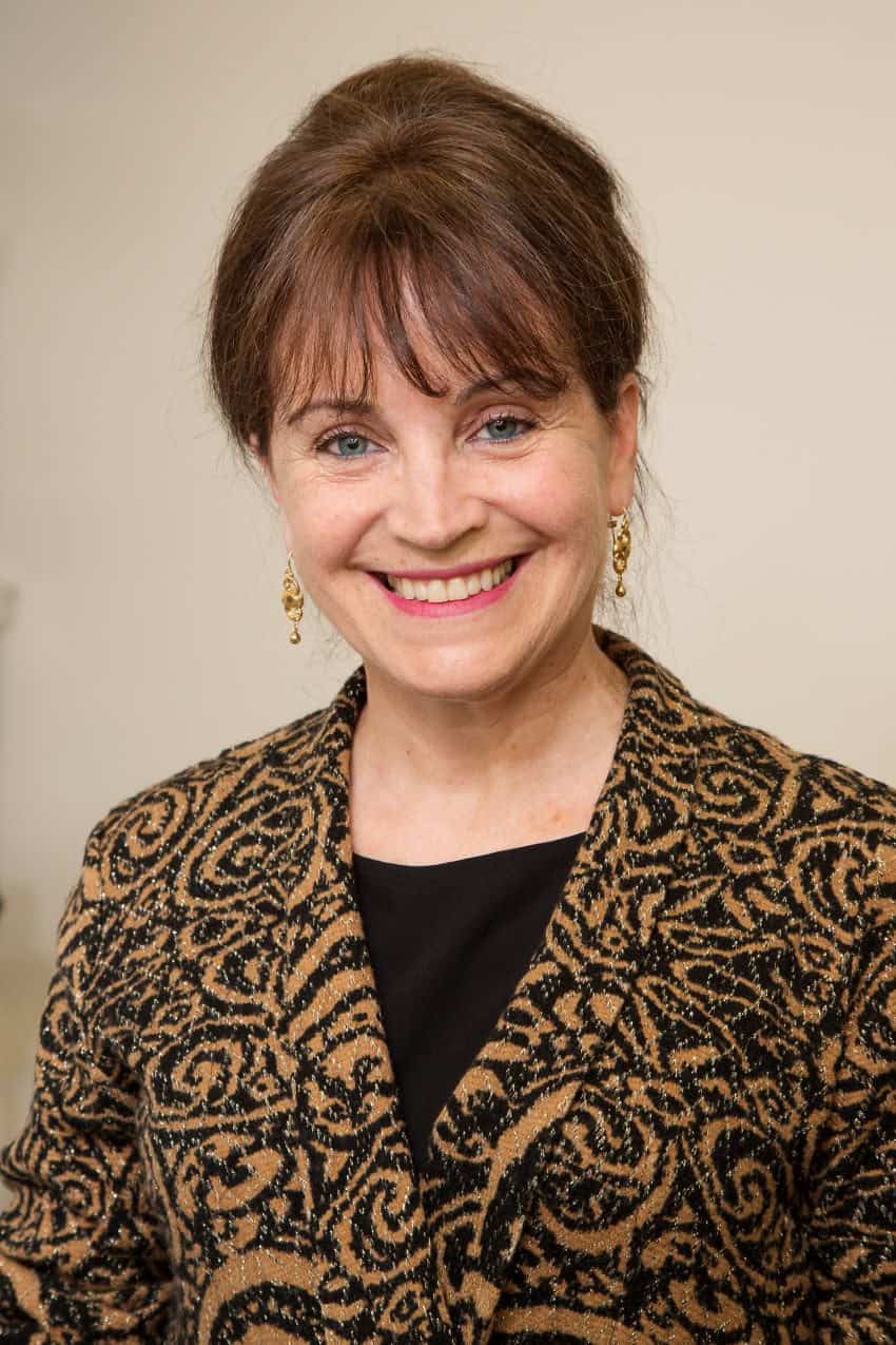 Headshot of a woman in a brown and black patterned jacket. She wears her hair up.