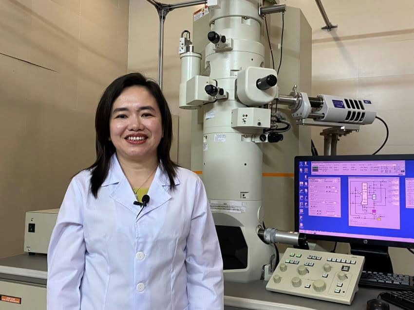 Woman wearing a lab coat with purple screen and equipment behind.