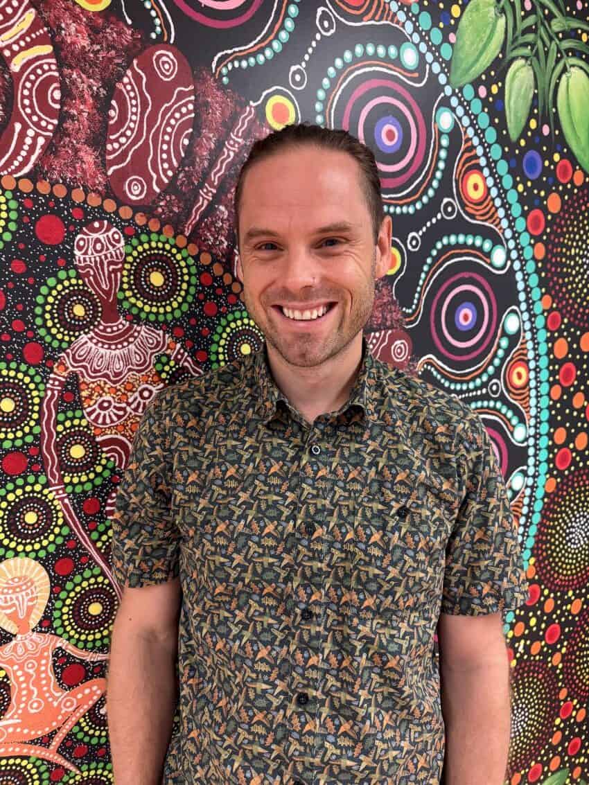 Head and shoulder photo of a man standing in front of a wall painted in colourful indigenous dot art design.