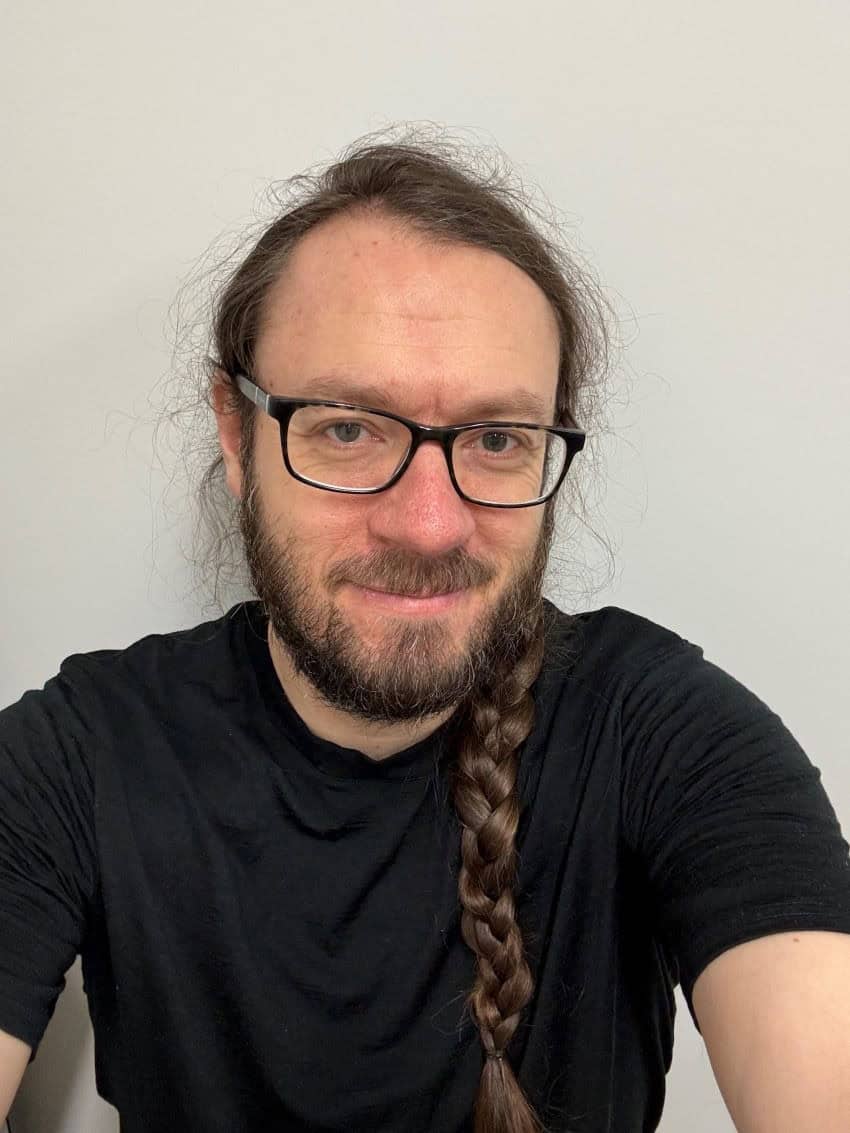 Head and shoulders shot of sam baron. Male with plaited hair and beard. Wearing glasses and a black t-shirt.
