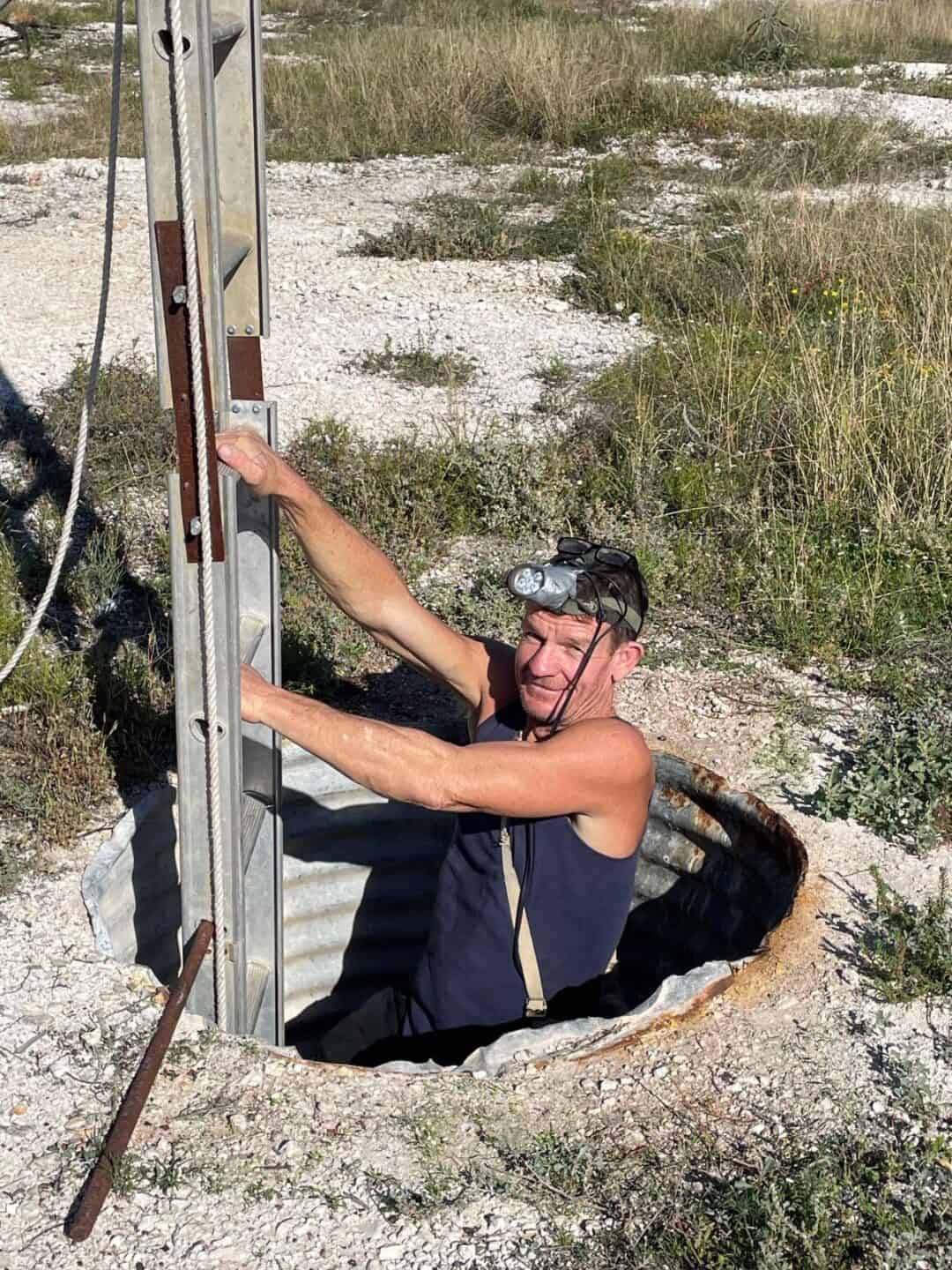 A man wearing a head torch climbs down a ladder into a hole in the ground.
