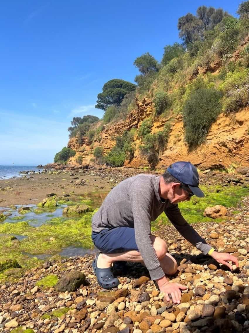 Man crouching on a rocky foreshore picking up a rock.