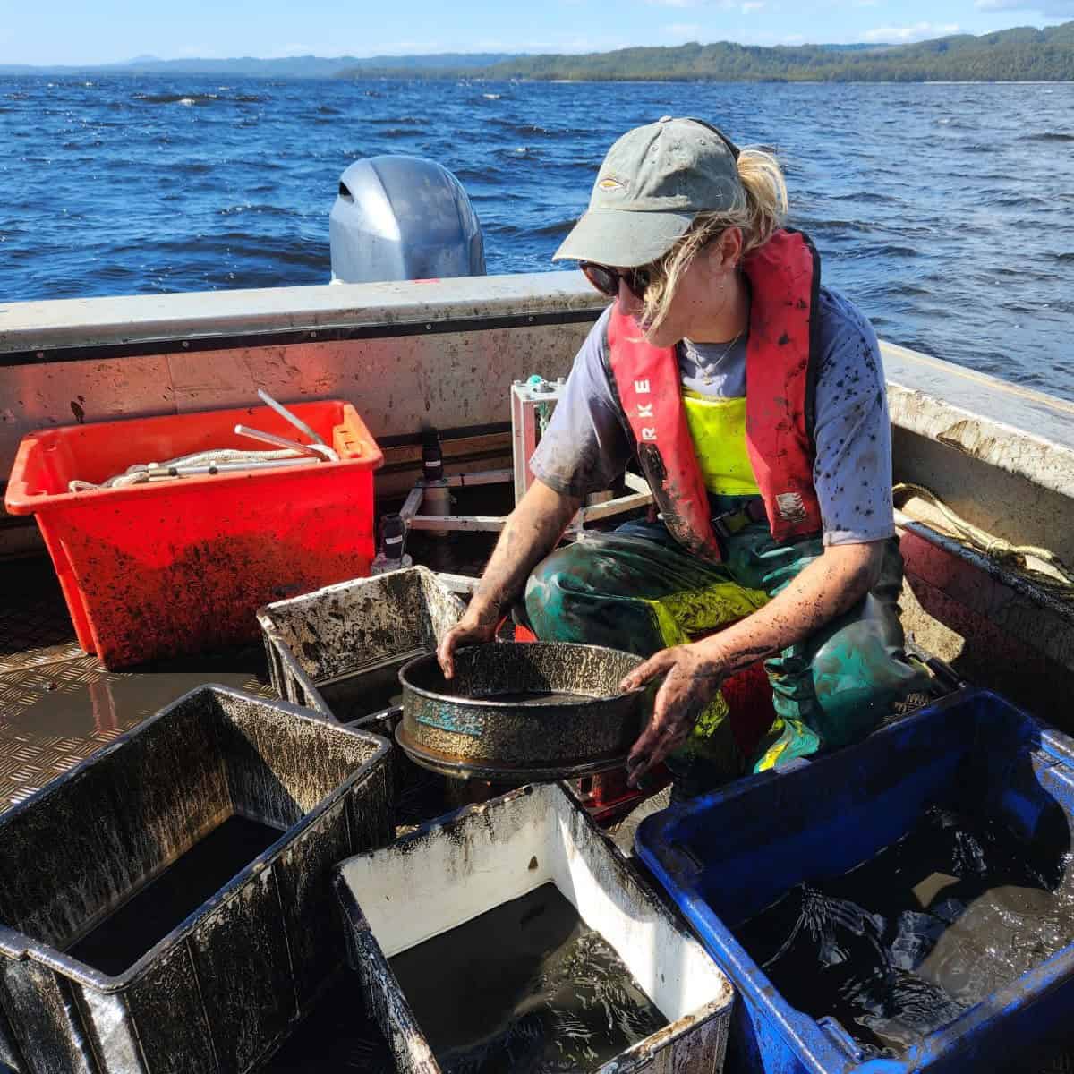 Woman crouching on a boat, looking at containers of black liquid. She is splashed with black muck.