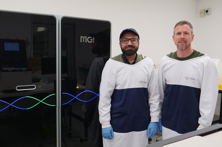 Two men wearing lab coats and gloves stand in front of a glossy black piece of technology the size of a large refrigerator