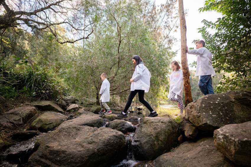 5 children wearing white lab coats climb over rocks in a creek