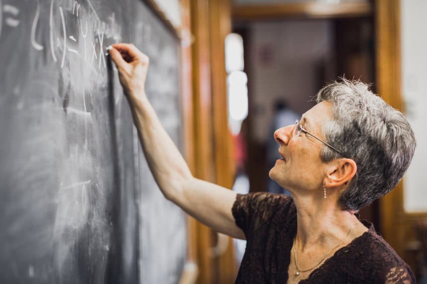 A woman with short grey hair and glasses works on a maths problem on a blackboard