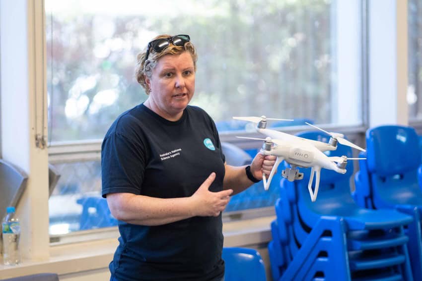 A woman wearing a black tshirt holds a white drone while speaking to an audience out of frame