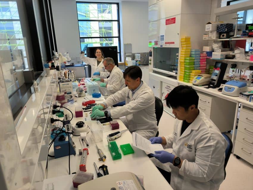 A group of researchers in lab coats prepare samples at a lab bench
