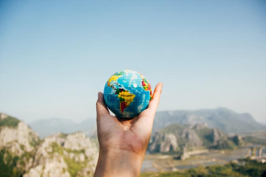 A hand holds a small globe in front of a scenery in the mountains