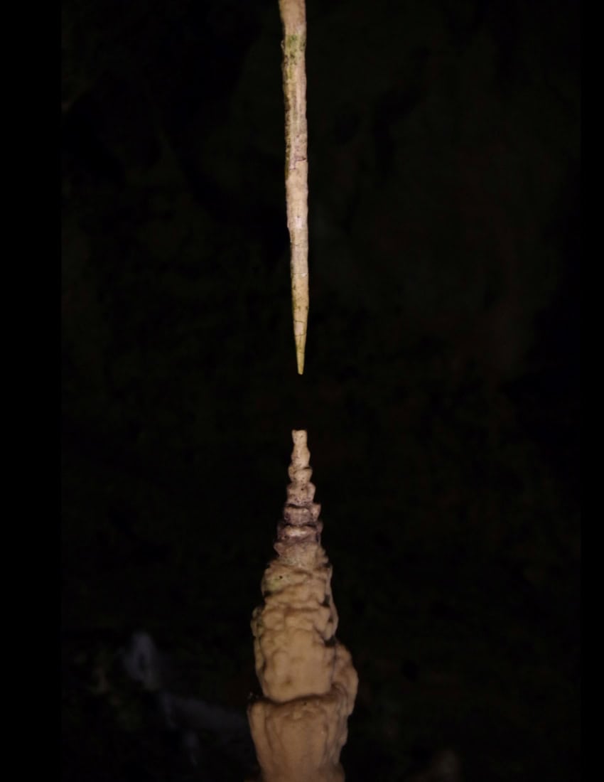A thin needle-like stalactite hangs above a cone-shaped stalagmite