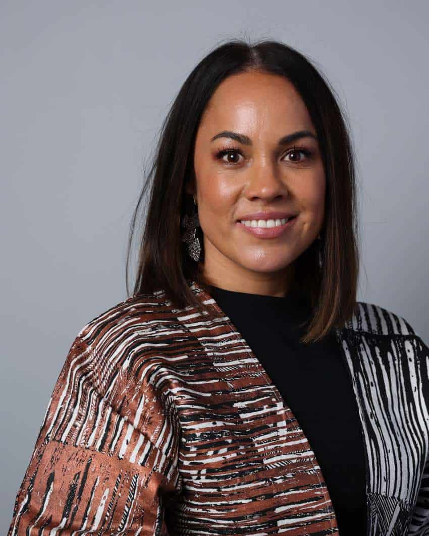 A photograph of a woman with shoulder-length, dark brown hair. She is wearing a brown, black, and white top.