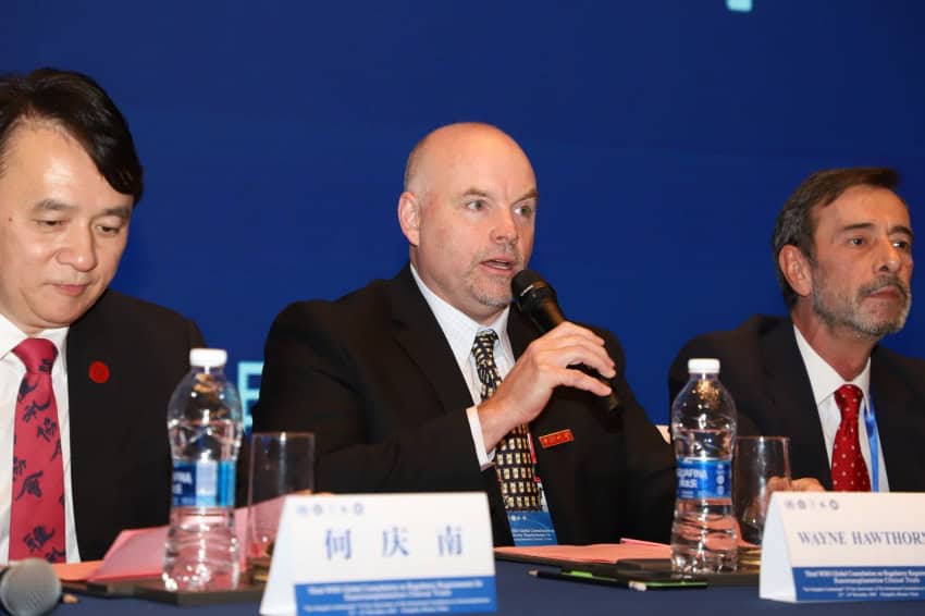 3 men sit at a panel table against a dark blue background. The central one is speaking into a microphone