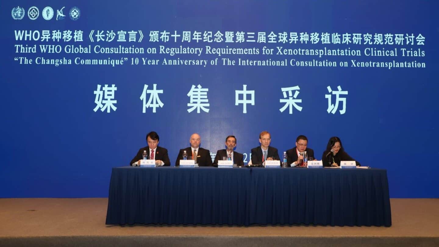 A panel of 5 men and one woman sits at a long table in from of a blue banner with white writing. It reads:third who global consultation on regulatory requirements for xenotransplantation clinical trials "the changsha communique" 10 year anniversary of the international consultation on xenotransplantation