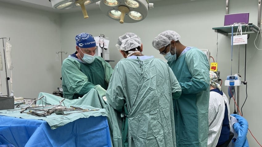 A team of surgeons in pale green ppe surrounds a person undergoing surgery