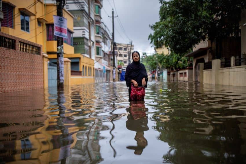 A photograph of a woman wearing dark coloured clothing and head covering and carrying a pink bag wading through thigh-high floodwater in the middle of a street.