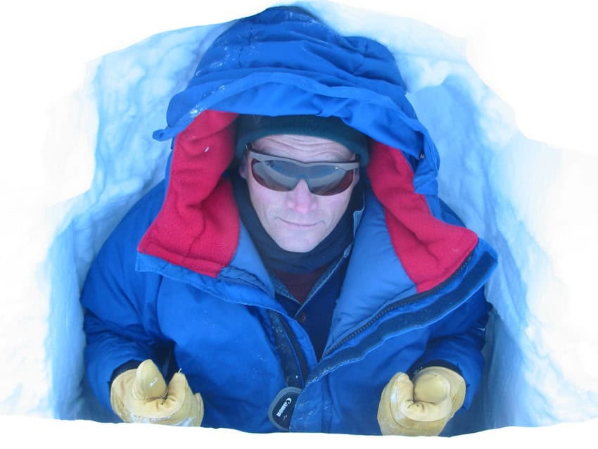 A photograph of a man in a big blue and red winter jacket. Black beanie, sunglasses, and yellow gloves. He is photographed in what appears to be sitting in a dug-out area of snow about large enough to fit one person.