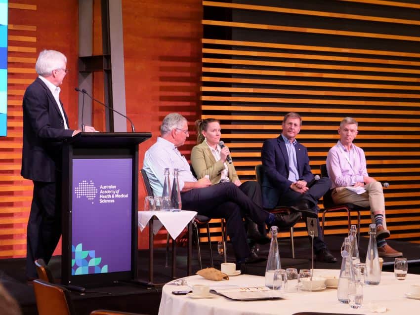 A photograph of a panel of 4 experts sitting on chairs next to a speaker's podium,