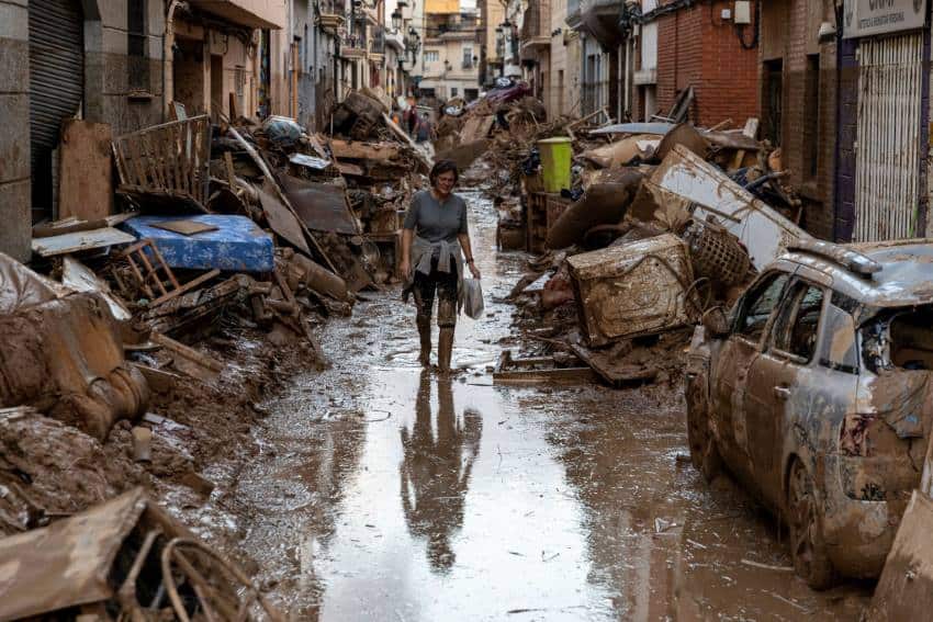 A photograph of a woman walking through a flooded street. It is covered in mud and there is detritus piled up on either side