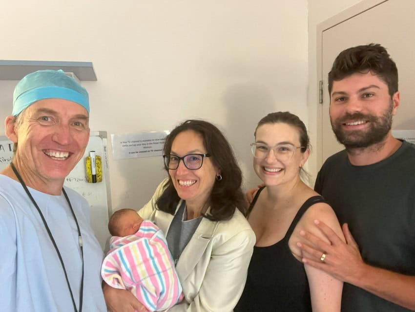 A photograph of 4 adults and a baby. On the left is a man dressed in blue scrubs, a woman with dark brown hair and glasses holding a newborn baby. On the left are a young couple, the woman is wearing a black tank top and glasses and the man is wearing a dark t-shirt.