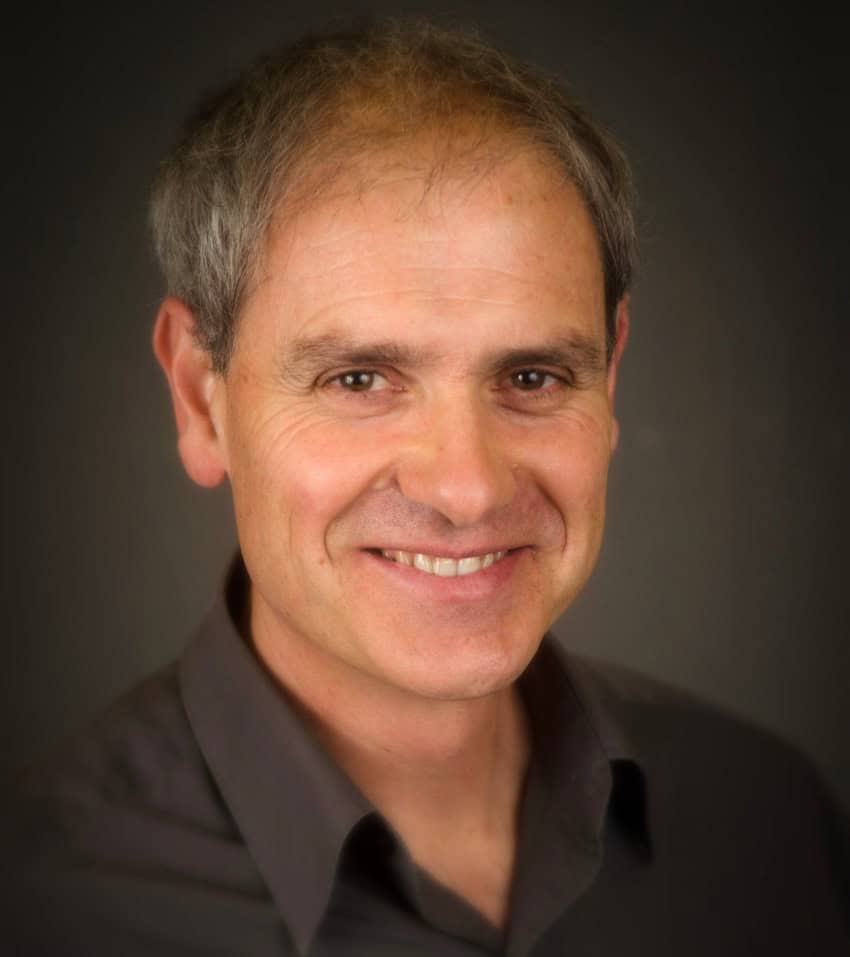 A photograph of a man smiling at the camera. He has greying hair and is wearing a dark coloured shirt.