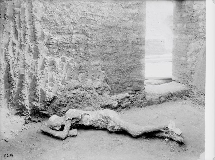 A black and white photograph of a plaster cast of a human body laying down on the ground, placed in the ruins of pompeii